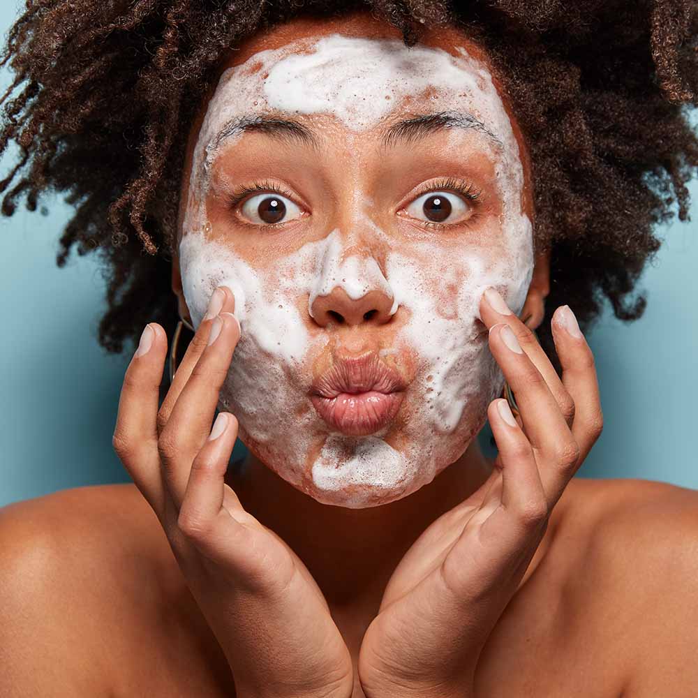 People, wellness, hygiene and skin problems concept. Lovely Afro American lady keeps lips folded, touches cheeks, has white foam on faces, washes with beauty gel, feels refreshed, has wide opened eyes